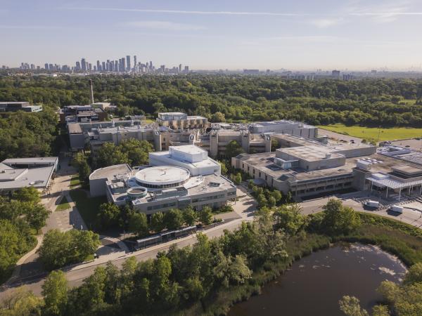 Aerial view of the U of T Mississauga campus surrounded by trees and a small pond, with modern buildings in the foreground and the Mississauga skyline in the background