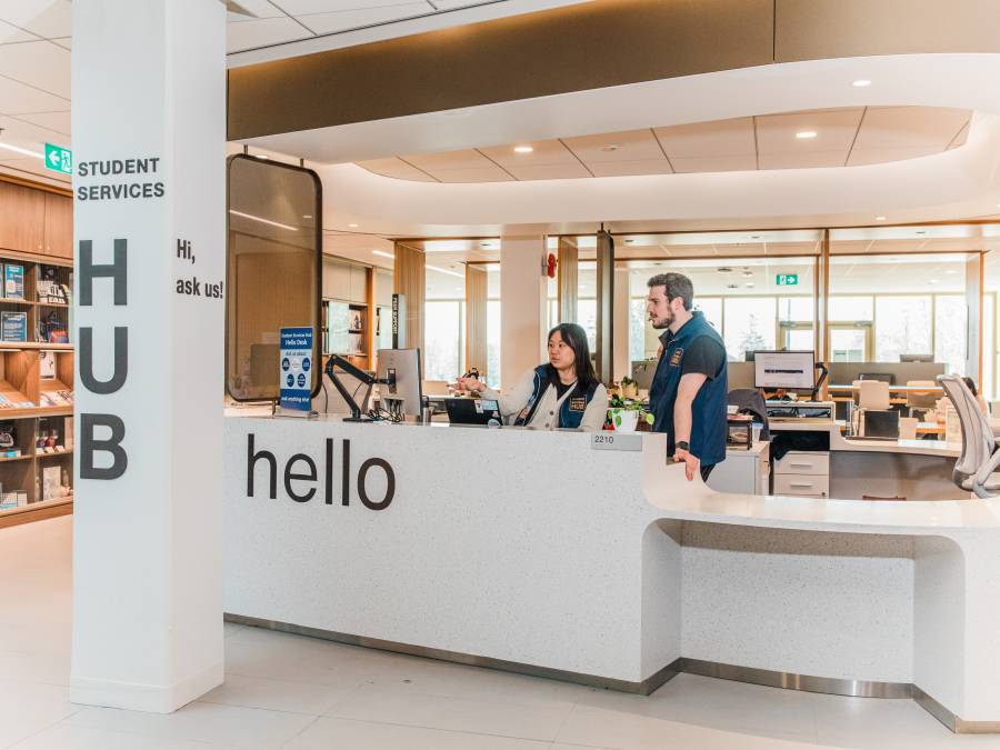 Two people speak at the Student Services Hub desk, with a “hello” sign and shelves in the background