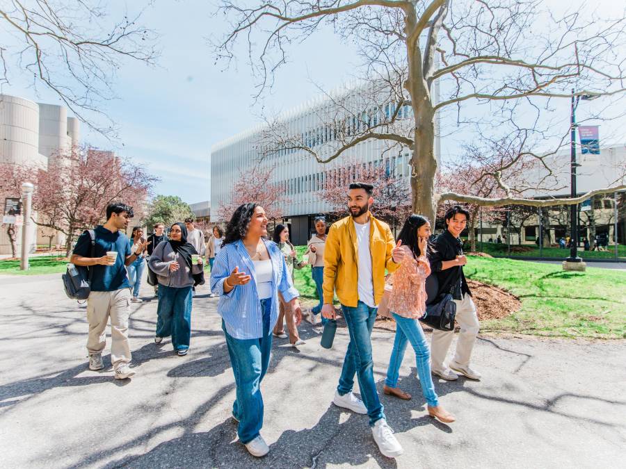 A group of prospective students walking together on a sunny day, chatting and smiling as they pass modern academic buildings and budding trees