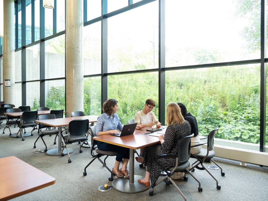 Students sitting at a table inside, next to a window-lined wall looking out lush greenery