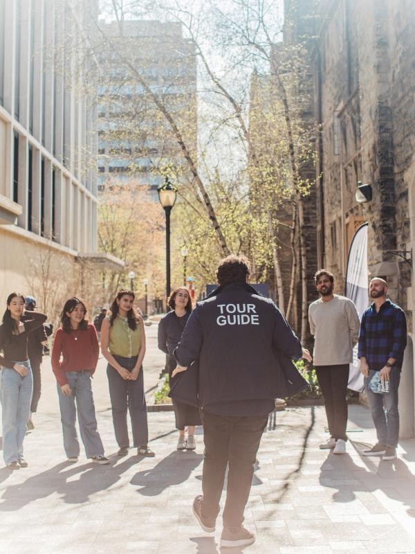 Campus tour guide wearing a navy jacket labeled “TOUR GUIDE” leads a group of high school–age prospective students down a sunlit pathway