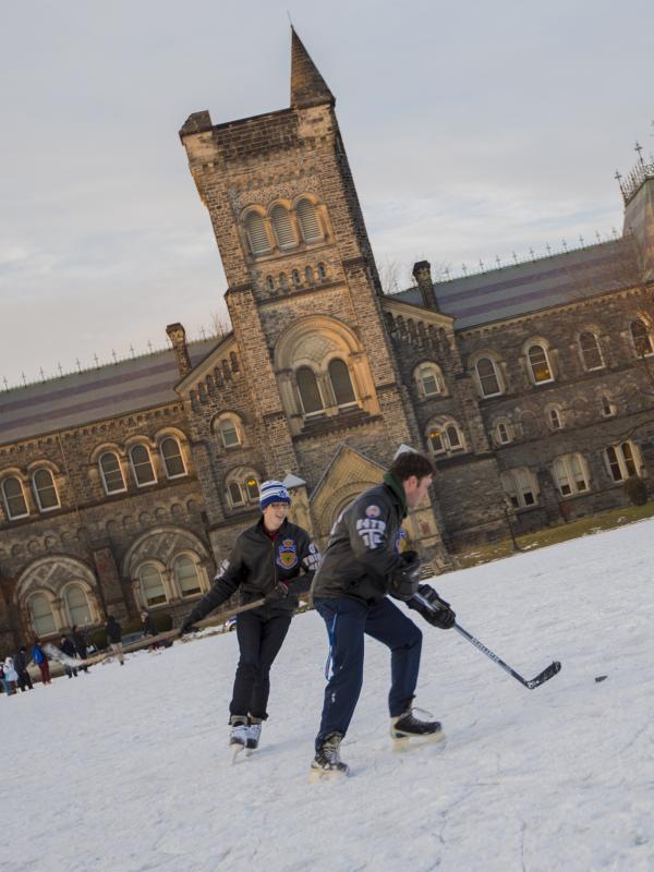 Two people playing ice hockey on a frozen Front Campus with University College in the background