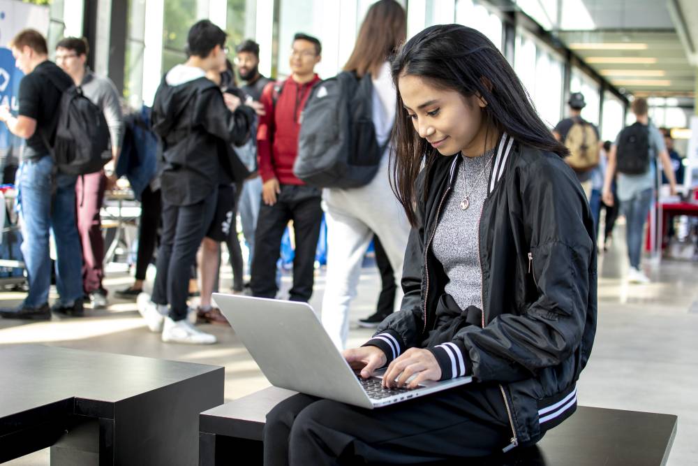 Student using laptop while sitting on bench in crowded hallway.