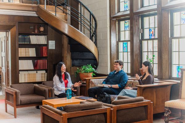 Three students sitting and talking in a wood-paneled study space with large stained-glass windows and a spiral staircase