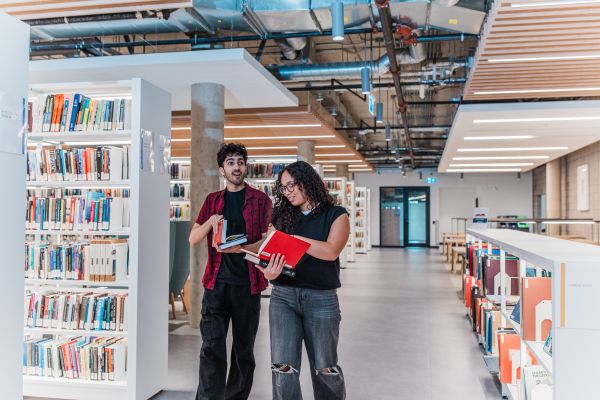 Two students standing next to each other, reading books in the library