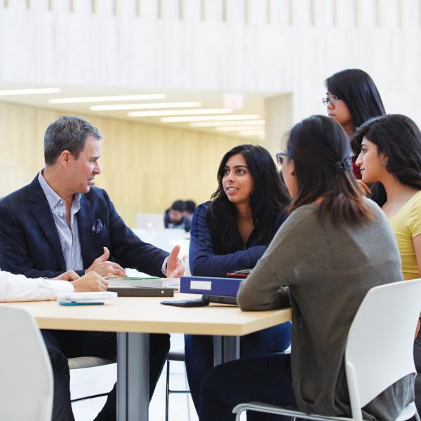 Students sitting around a table speaking with a professor inside a bright building atrium