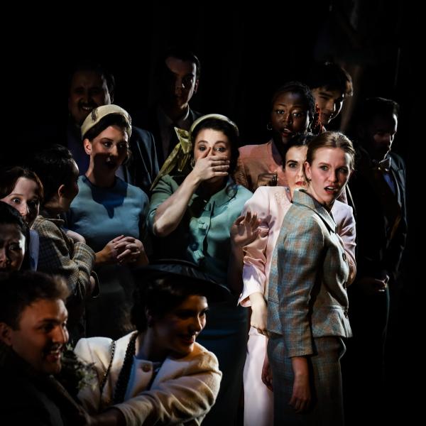 A group of actors on stage in dramatic lighting, dressed in vintage-style costumes, reacting with shock during a theatrical performance