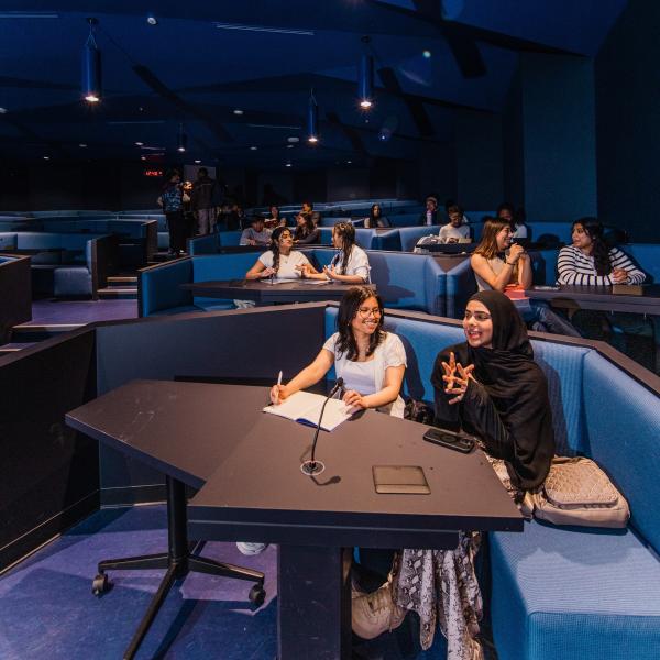 Students seated in groups in a dark lecture hall engaging in discussion