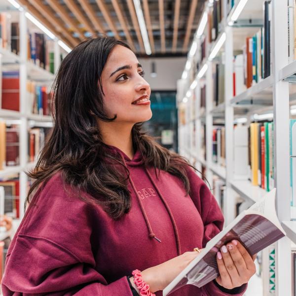 Student standing in a library aisle, smiling and holding an open book, wearing a maroon sweater